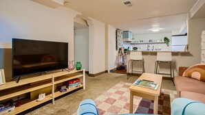 A living room with a TV, a coffee table, and a sofa. There is a kitchen in the background with a dining area.