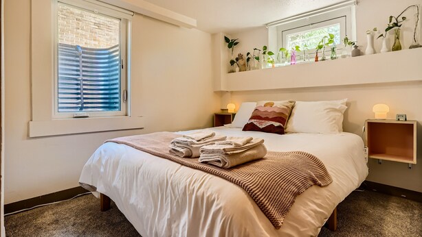 A neatly made bed with a folded towel, a bedside table with a lamp, and a window with blinds.