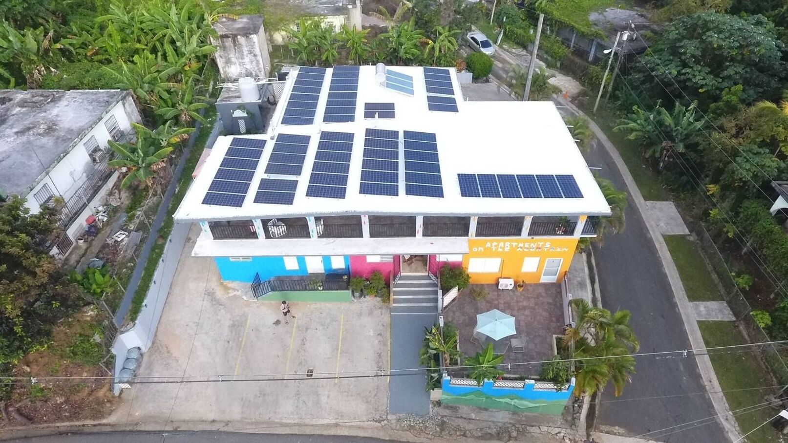 A building with solar panels on the roof, a parking area, and a sign indicating "APARTMENTS on the 2nd floor."