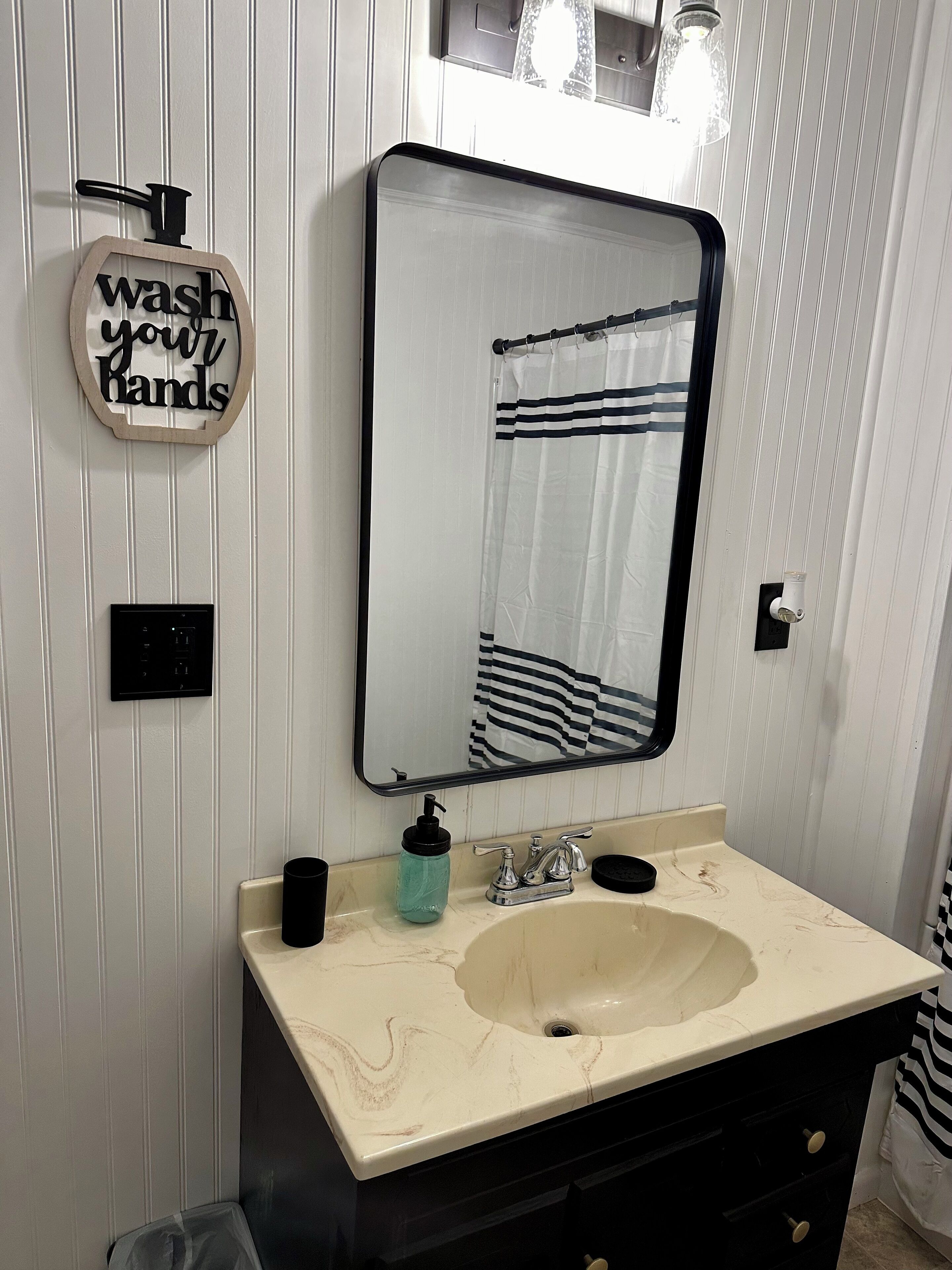 Une salle de bain avec un meuble-lavabo blanc, un évier, un miroir, un distributeur de savon et un panneau indiquant : « Lavez-vous les mains ».