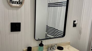 Une salle de bain avec un meuble-lavabo blanc, un évier, un miroir, un distributeur de savon et un panneau indiquant : « Lavez-vous les mains ».