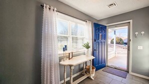 Un pasillo con una puerta azul, una mesa blanca y una cesta, y una ventana con cortinas blancas.