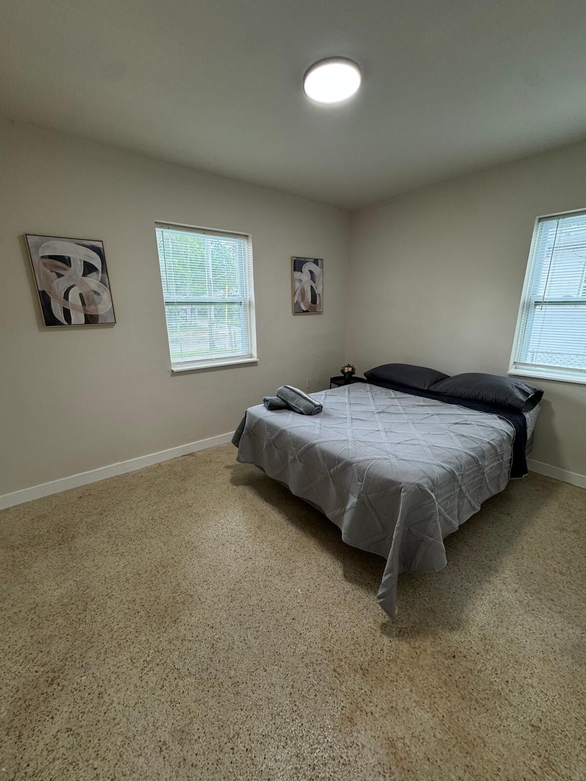 A bedroom with a bed, two windows with blinds, and two abstract paintings on the wall.
