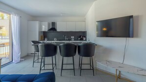 A modern kitchen with white cabinets, a black range hood, and a dining area with three black bar stools. A flat-screen TV is mounted on the wall above the counter.
