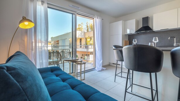 A modern apartment with a blue sofa, a small dining area with two chairs, a kitchen with white cabinets, and a balcony with a view of buildings.
