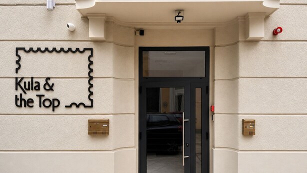 L’entrée d’un hôtel, avec une porte noire, une caméra de surveillance au-dessus et une enseigne indiquant « Kula & the Top ».