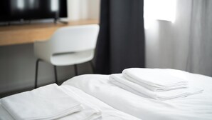 Un lit bien fait, avec des draps blancs soigneusement pliés, une chaise blanche et un téléviseur en arrière-plan.