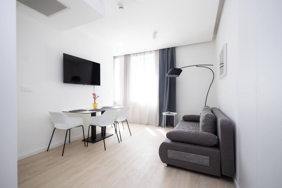 Una sala de estar moderna con un televisor de pantalla plana, un sofá gris, un comedor con una mesa y sillas, y un ventanal con cortinas.