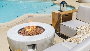 A poolside area with a circular stone fire pit, wicker furniture, and a lantern.