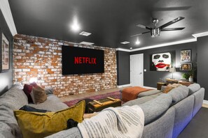 A living room with a brick wall, a large flat-screen TV, a ceiling fan, and a grey sofa. - Iconic Vegas Villa : Heated Pool| Movie Room (Las Vegas)