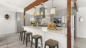 A modern kitchen with a large island, bar stools, hanging pendant lights, and a rustic wooden beam.