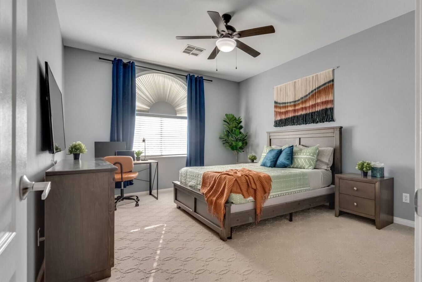 A bedroom with a bed, a desk, a chair, a dresser, a ceiling fan, and a window with curtains.