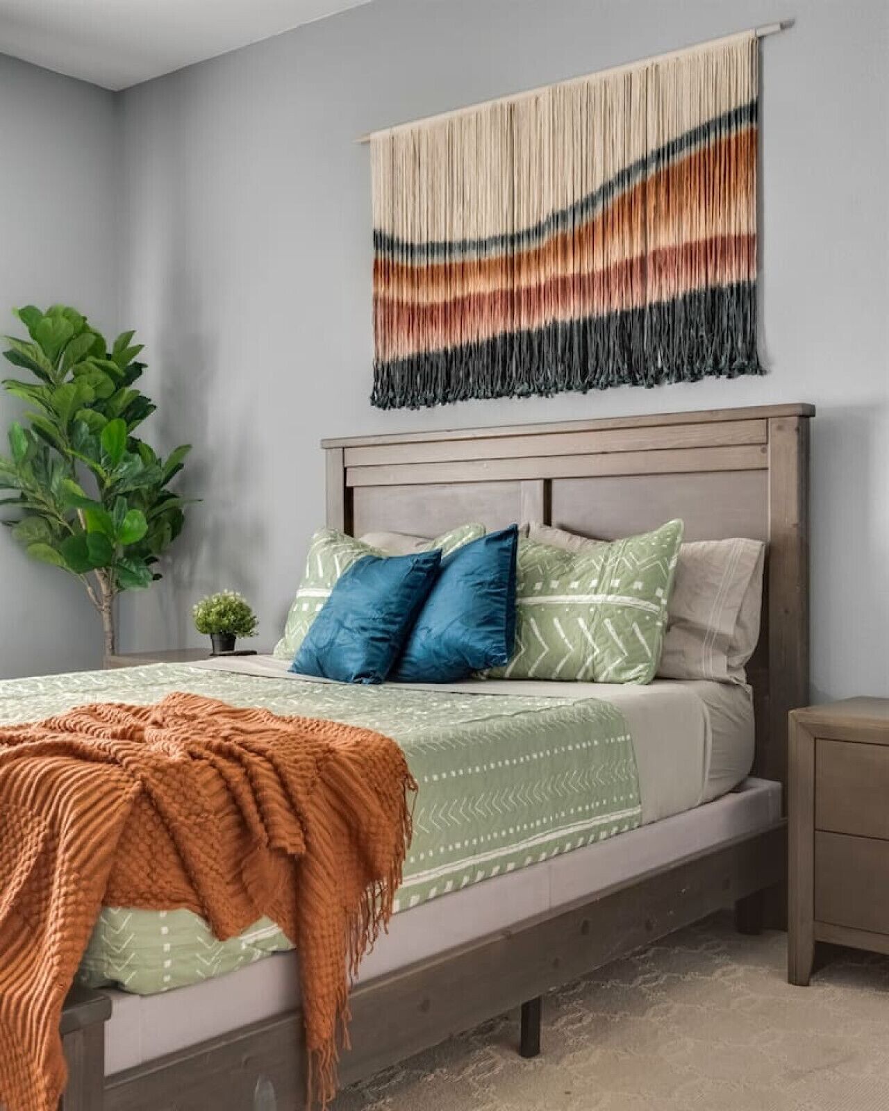 A bedroom with a bed, pillows, a blanket, a wall hanging, and a plant.
