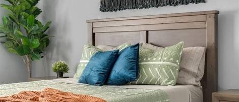 A bedroom with a bed, pillows, a blanket, a wall hanging, and a plant.