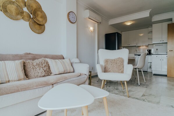 Una sala de estar moderna con un sofá, un sillón y una mesa de centro. Se observa una cocina al fondo con armarios y un refrigerador.