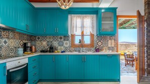 A kitchen with teal cabinets, a patterned backsplash, a window with white curtains, and a view of a patio. - Holiday Home 'Archontiko Keli' with Mountain View, Wi-Fi and Air Conditioning (Katakilos)