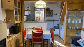 Ett kompakt kök med träskåp, ett matbord med stolar, ett litet kylskåp och ett vitrinskåp i glas där köksredskap förvaras.