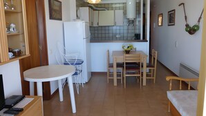 A compact living space with a kitchenette, a dining area with a table and chairs, a small seating area with a bench, and a television on a stand. - Apartment 'Platja Mar 2' with Sea View and Balcony (L'Escala)