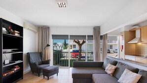A modern living room with a sofa, armchair, and bookshelf, a balcony with a view of buildings and parked cars.