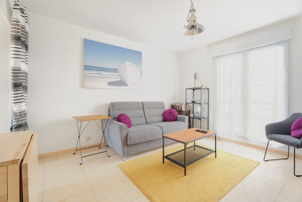 A modern living room with a sofa, coffee table, and a painting of a boat on the wall. - Apartment 'Le Saint Martin' with Sea View, Balcony and Wi-Fi (Bréhal)