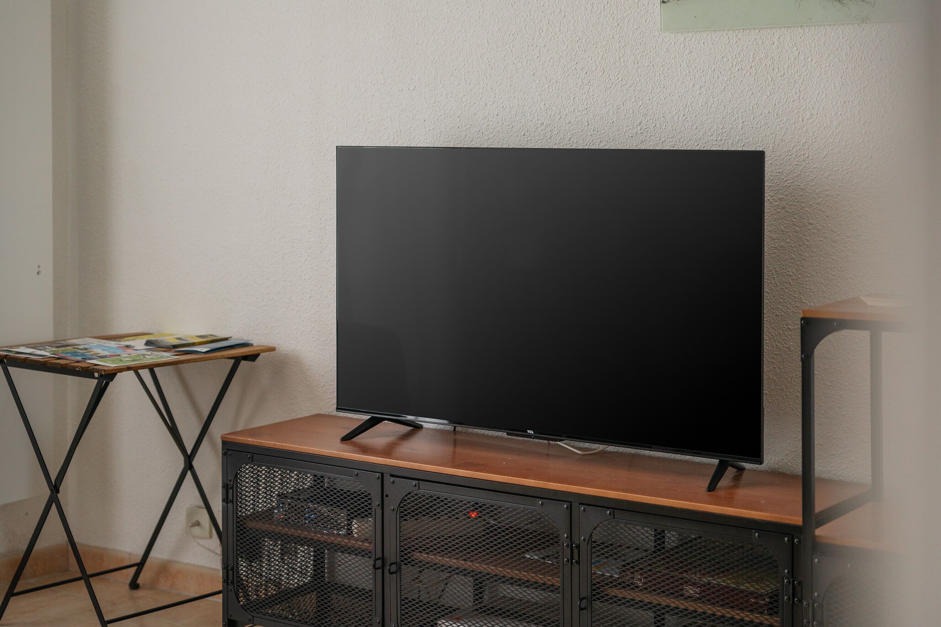 Un téléviseur posé sur un meuble en bois avec un cadre en métal, une petite table avec des magazines et un haut-parleur fixé au mur.