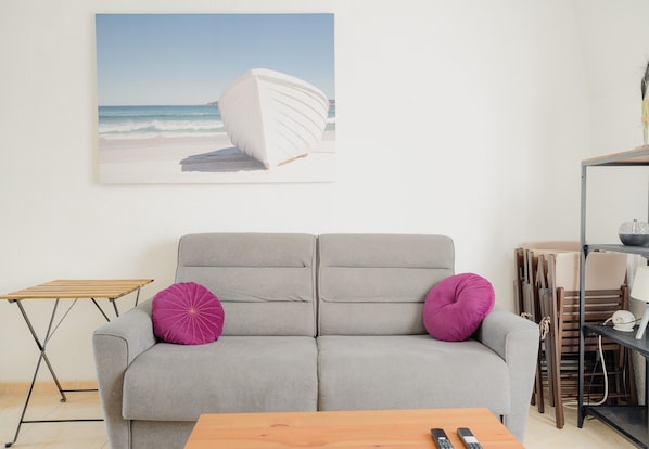 Un salon avec un canapé gris, deux coussins violets, une table basse en bois, un tableau représentant un bateau sur une plage et une étagère en métal sur laquelle sont rangées des chaises pliantes.