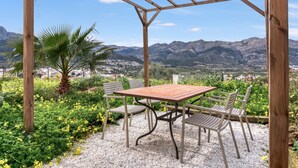 A wooden pergola with a table and chairs, surrounded by greenery and a view of mountains.