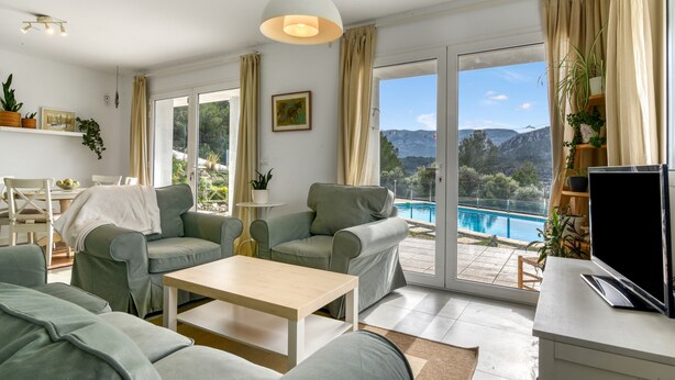 A modern living room with a pool view, green sofas, a wooden coffee table, and a TV on a white cabinet.