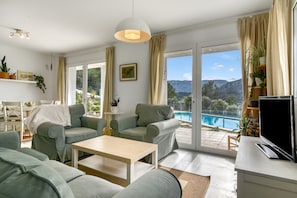 A modern living room with a pool view, green sofas, a wooden coffee table, and a TV on a white cabinet. - VillaSol – Peaceful House with Private Pool and Mountain Views (l'Aspre)