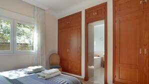 Ein Schlafzimmer mit Bett, Holzschrank, Stuhl und angrenzendem Badezimmer, das durch eine geöffnete Tür zu sehen ist.