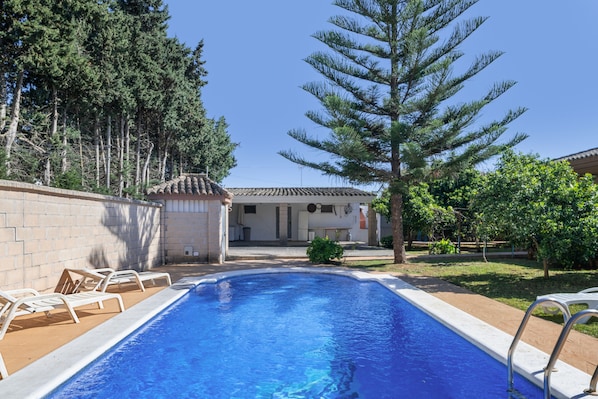 A swimming pool with lounge chairs, a building with a tiled roof, and surrounding trees.