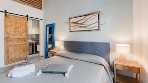 Un dormitorio con cama, armario de madera, puerta azul, obra de arte colgada en la pared y lámpara de noche.