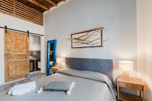 A bedroom with a bed, a wooden wardrobe, a blue door, a wall-mounted artwork, and a bedside lamp.
