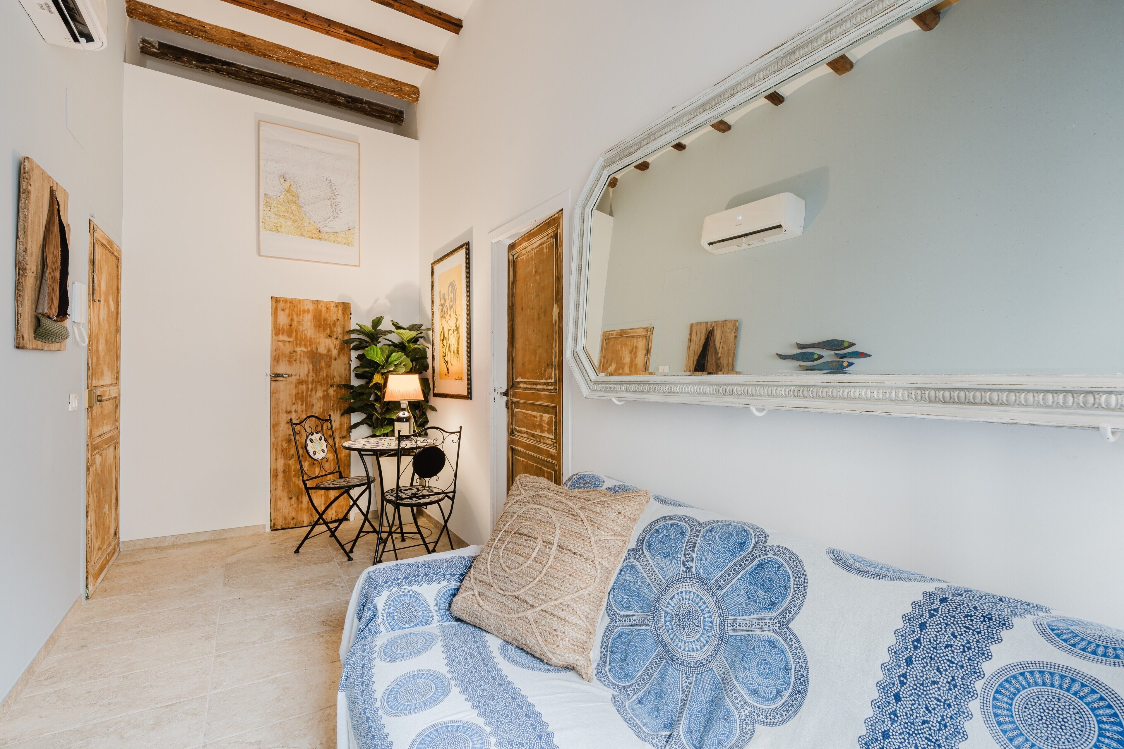 A bedroom with a bed, a mirror, a wooden door, a small table with a lamp, and a chair.