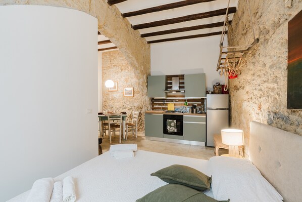 A bedroom with a bed, pillows, a bedside table with a lamp, a kitchen area with a stove, and a dining table with chairs.