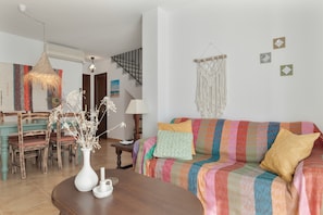 A living room with a sofa, a wooden dining table, chairs, a vase with dried flowers, and a hanging macrame piece.