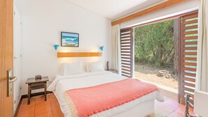 A bedroom with a bed, a nightstand, a window with blinds, and a view of trees. - Apartment 'Cerca Do Altinho' with Private Pool, Private Terrace and Wi-Fi (Vila Nova de Milfontes)