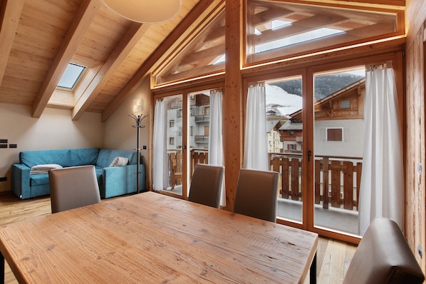 A wooden interior with a dining table, chairs, a sofa, and a view of a mountain landscape through large windows.