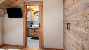 Une pièce avec un plafond en bois, un téléviseur fixé au mur, une armoire en bois et une salle de bain visible à travers une porte ouverte.