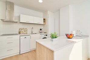 Une cuisine moderne avec des meubles blancs, un lave-vaisselle, un lave-linge et un plan de travail agrémenté d’une plante et d’un panier de fruits.
