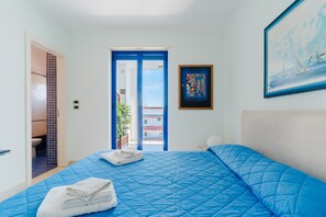 Une chambre à coucher avec une couvre-lit matelassée bleue, une tête de lit blanche, un tableau encadré et une vue sur un balcon extérieur, visible à travers une porte.