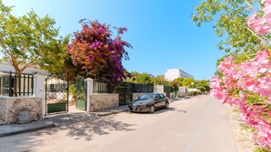 Vista de una calle con un coche estacionado junto a una entrada protegida por un portón, un muro de piedra y un árbol con flores rosadas.
