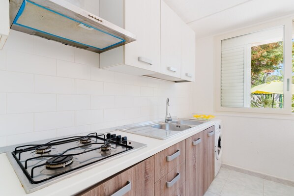 Une cuisine avec des meubles blancs, une cuisinière à gaz, un évier et une fenêtre donnant sur des arbres.