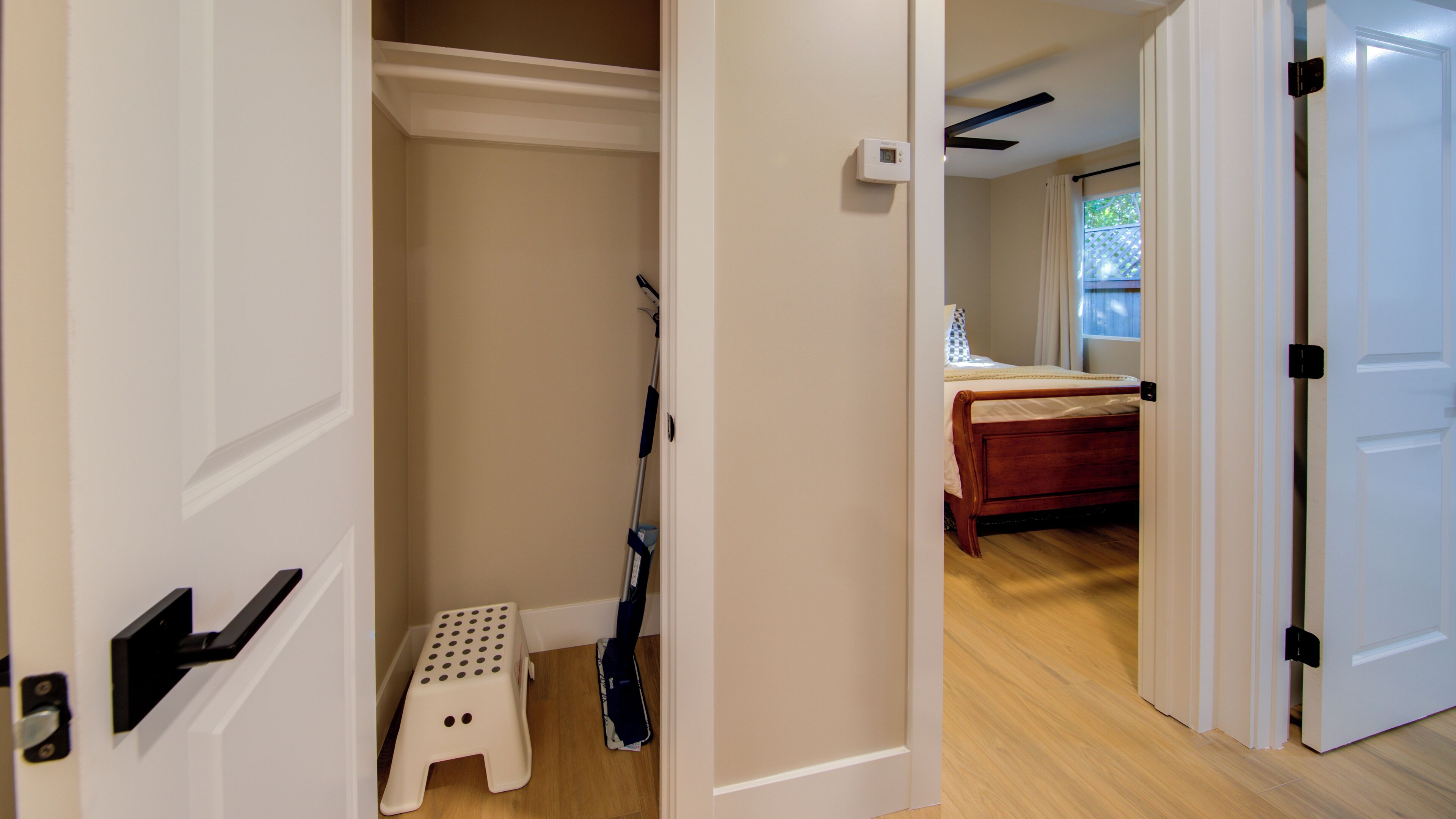 A hallway with a wooden floor, a white door, and a step leading to a room with a bed and a window.