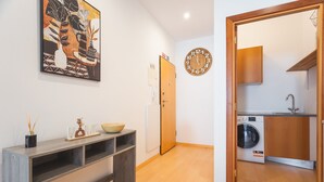 Un couloir doté d’un plancher en bois, d’une horloge murale, d’une porte, d’une console sur laquelle sont posés un bol et des objets décoratifs, ainsi qu’un coin buanderie avec un lave-linge.
