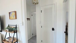 A hallway with a white door, a wall-mounted TV, a shelf with books and plants, and a hanging light fixture.
