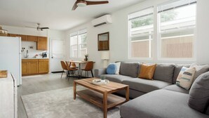 A spacious living area with a gray sofa, wooden coffee table, and a dining table with chairs. There is a ceiling fan, large windows, and an air conditioning unit.