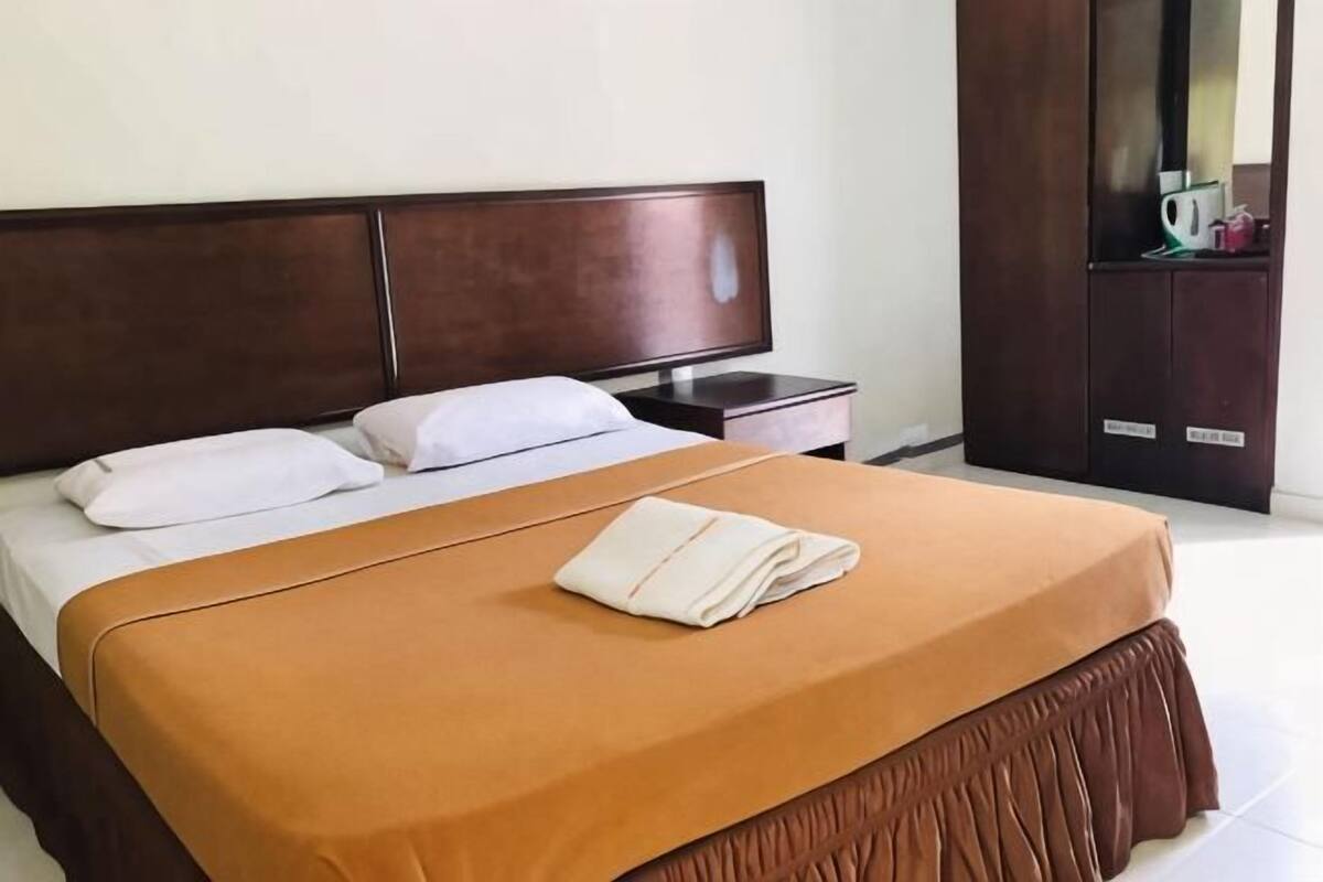 Un letto rifatto con copripiumino marrone e cuscini bianchi, una testiera in legno, un comodino con una lampada e un'armadio scuro in legno con uno specchio.