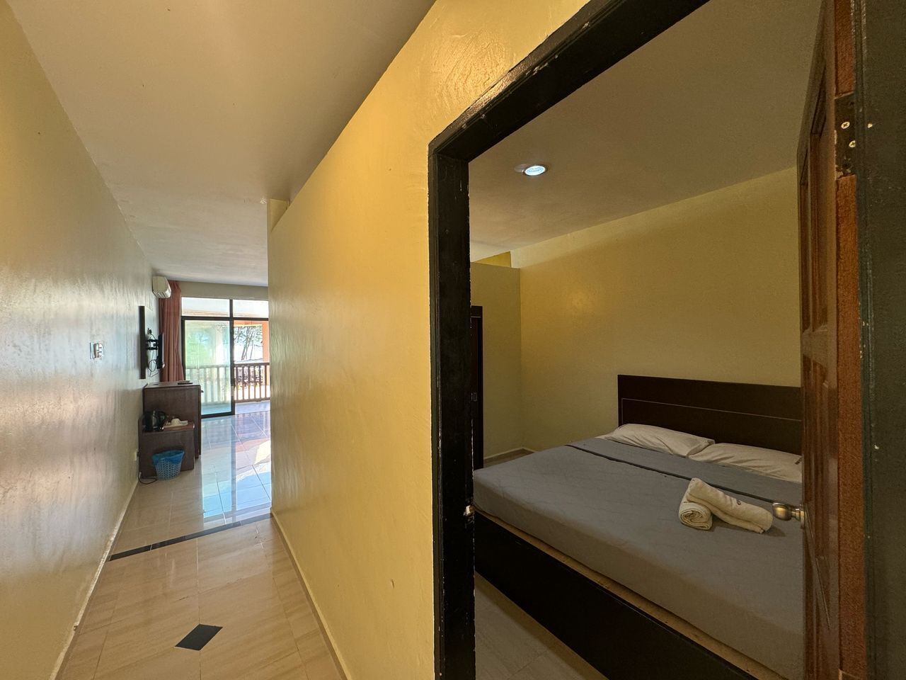 A narrow hallway leading to a room with a bed, a small desk, and a window.