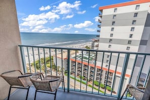 A balcony with wicker chairs, a view of the beach, and a hotel building with multiple floors.